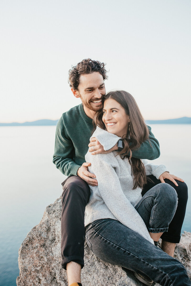 Couple embrace on rock on mountaintop overlooking Lake Tahoe