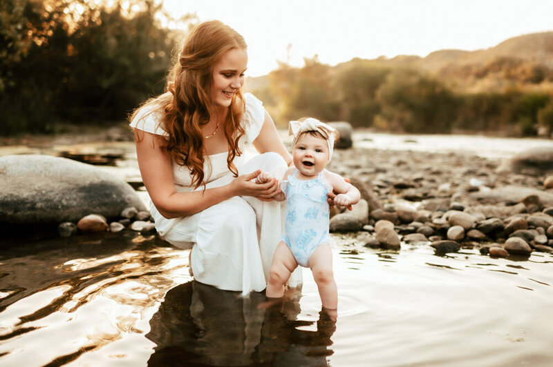 Mom squats down in a puddle with her baby girl while the sun sets behind them.