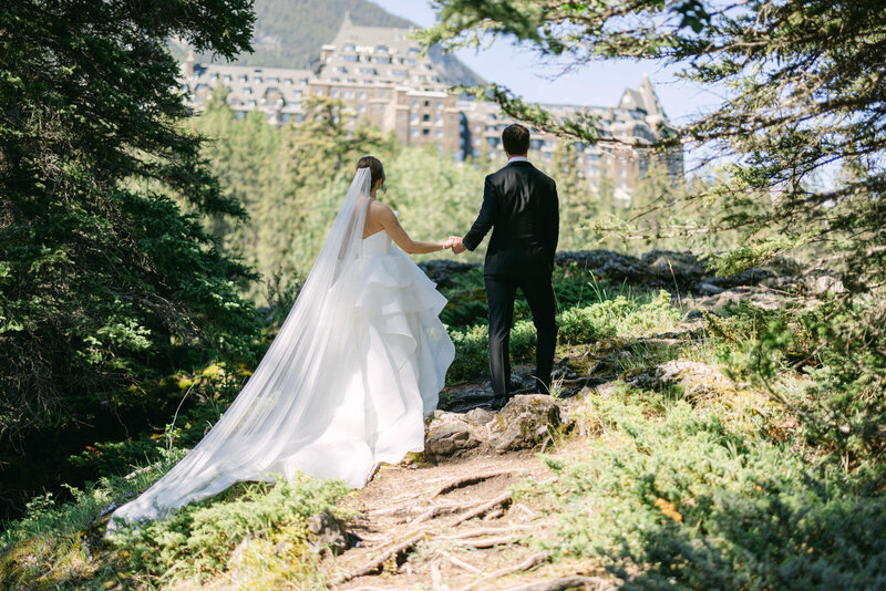 Bride and groom walking up a path toward the Fairmont Banff Springs Hotel surrounded by green trees and summer foliage in Banff National Park captured as a scenic wedding portrait
