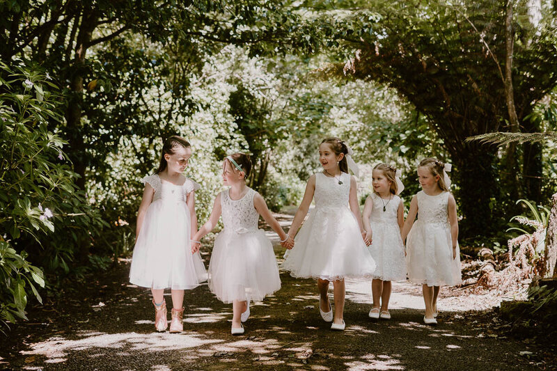 Flower girls holding hands, wearing white dresses, looking at each other candidly
