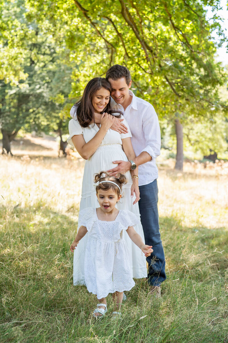 A family of three playing in the park during a photo shoot. The parents embrace while the little girl dances around them.