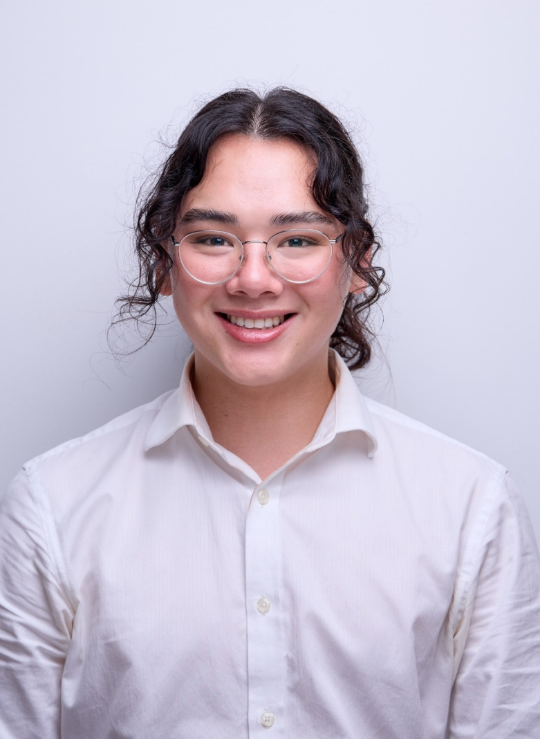 A close-up portrait of a person smiling. The person is wearing a shirt and the background seems to be a wall.