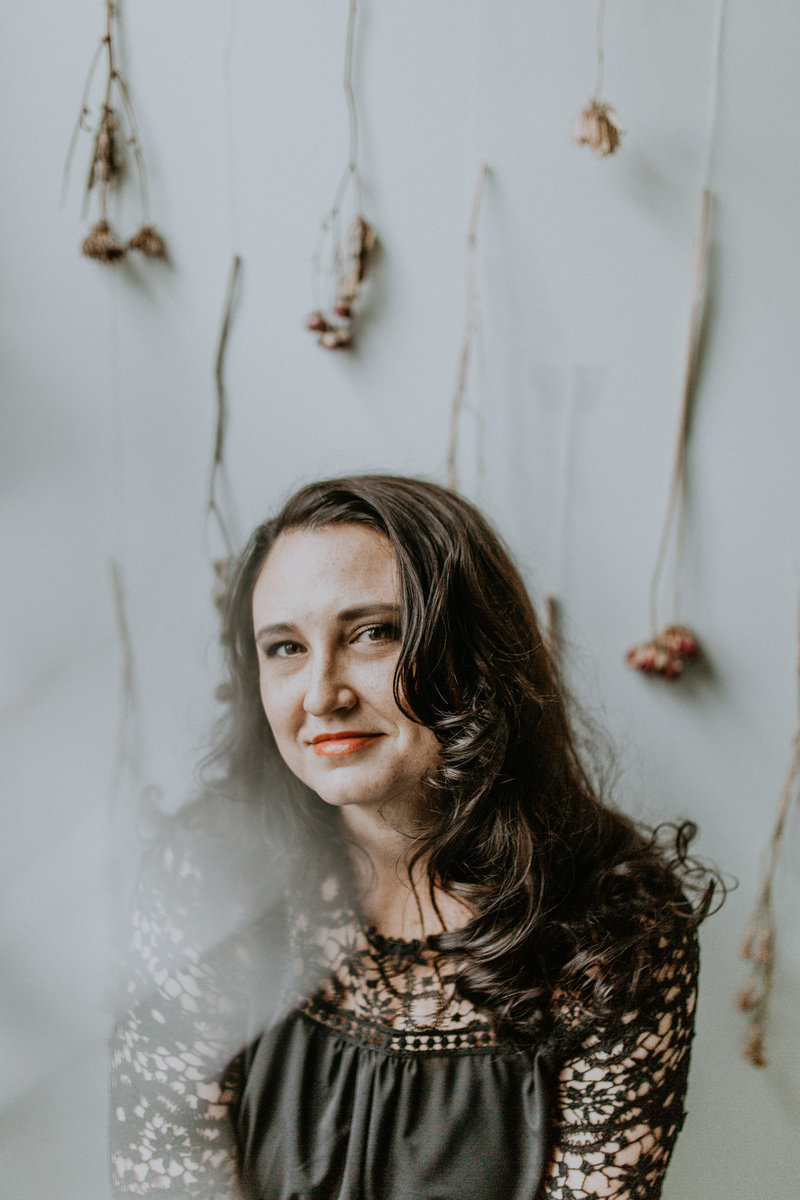 woman with dark hair sits in front of a camera  with flowers against a white wall in the background