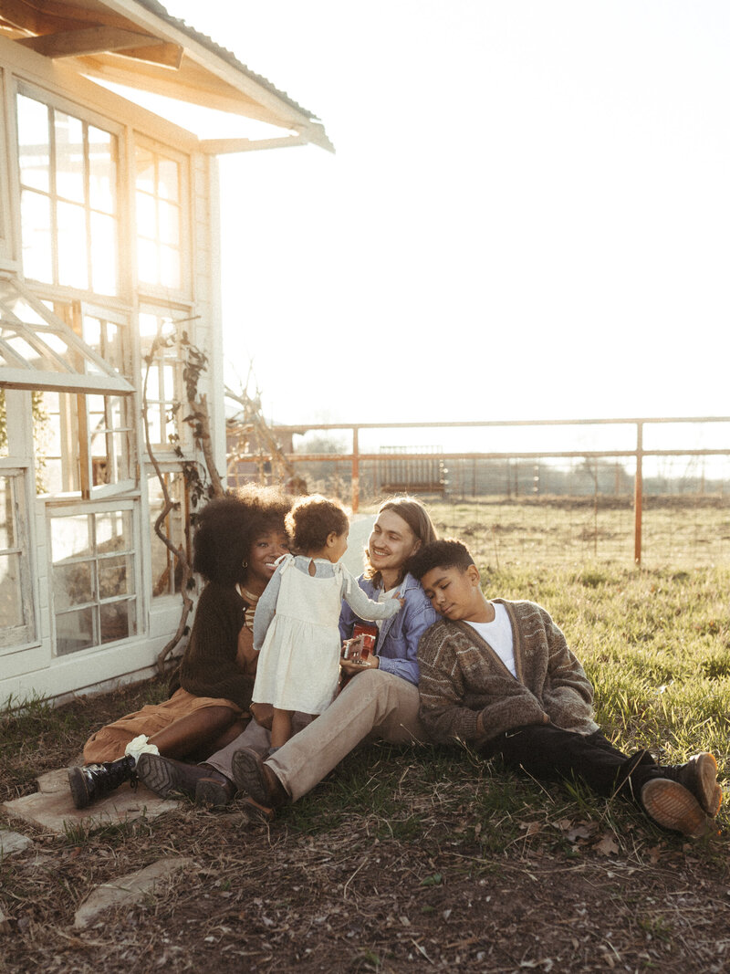 A family of four sit in a patch of shade while the sun shines through the glass shack beside them. 