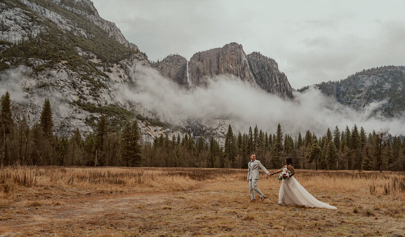 bride and groom walking across a meadow with yosemite falls in the background and fog hanging over the yosemite valley