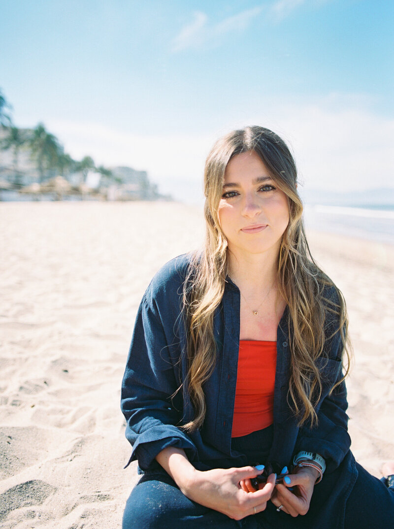 Photographer Little Rock Bailey Feeler sits on the sand of a Puerto Vallartan beach in a blue and red outfit.