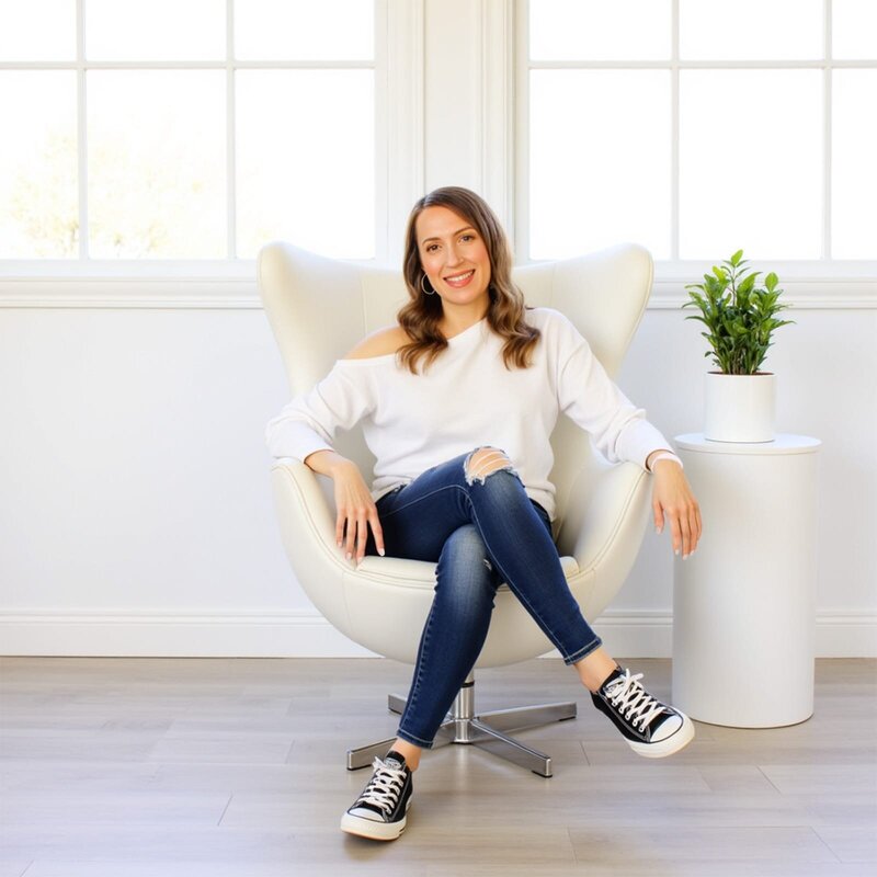 Stacey from Xanthe Bookkeeping smiling in a white off-the-shoulder sweatshirt and jeans while seated in a modern white egg chair with a green plant beside her, featured in the Contact page info section.