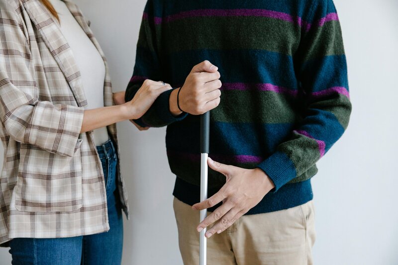 A person holds the arm of someone using a white cane, walking together indoors.
