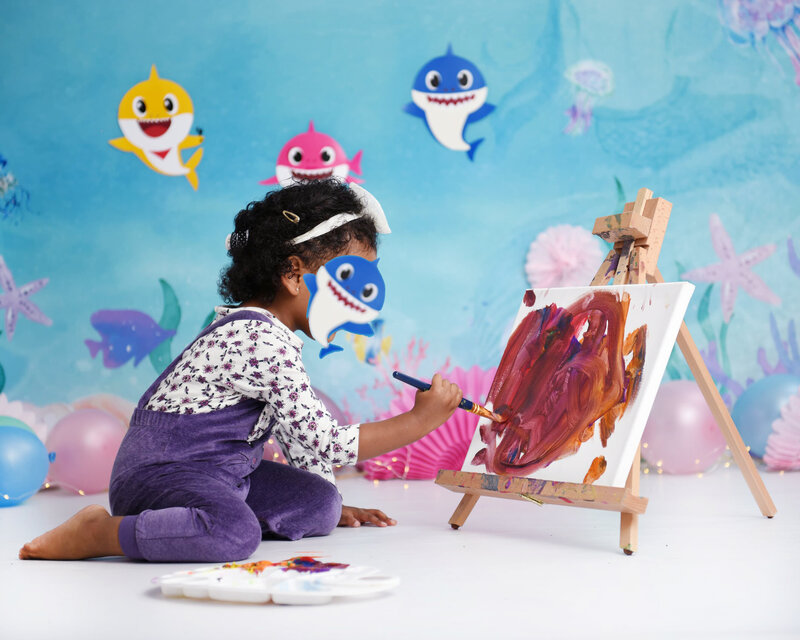 A toddler girl painting on a canvas during an underwater-themed photo shoot, featuring Baby Shark characters and ocean-inspired decor in the background.