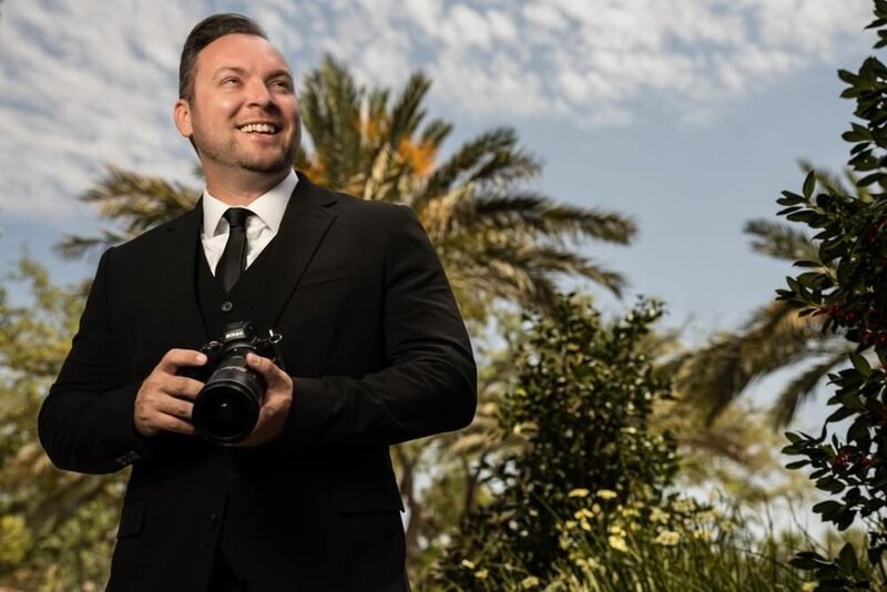 Man-in-a-suit-holding-a-camera-smiling-under-a-sunny-sky-with-palm-trees-in-the-background