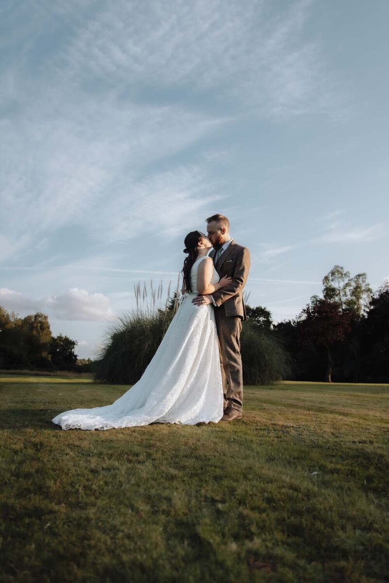 Bride & Groom kiss in field her dress trails behind