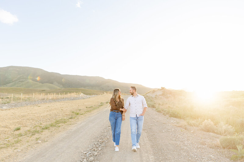 A couple walk closely down a dirt road as the sun sets behind them. They smile at each other while they hold hands.