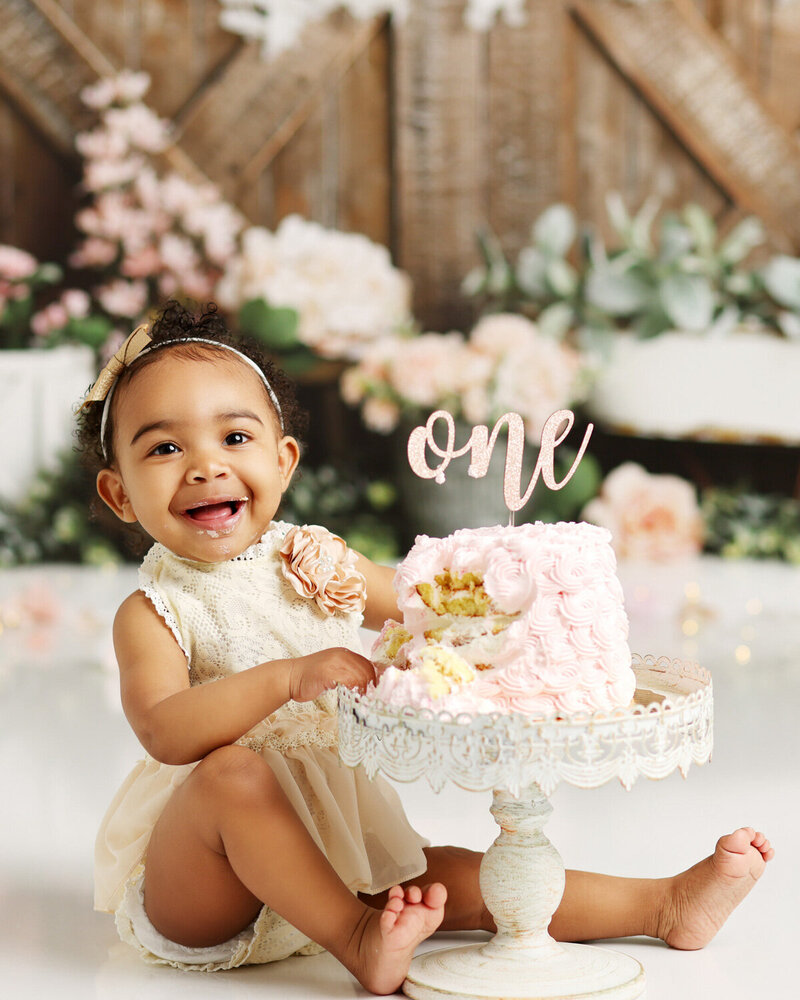 A joyful baby girl enjoying her first birthday cake smash. She sits beside a partially eaten pink cake with a "one" topper, surrounded by soft floral and rustic decor.
