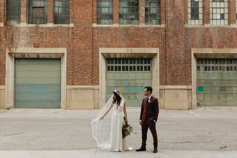 A bride and groom stand in front of a brick building, her veil flowing beautifully in the wind as they share a timeless moment.