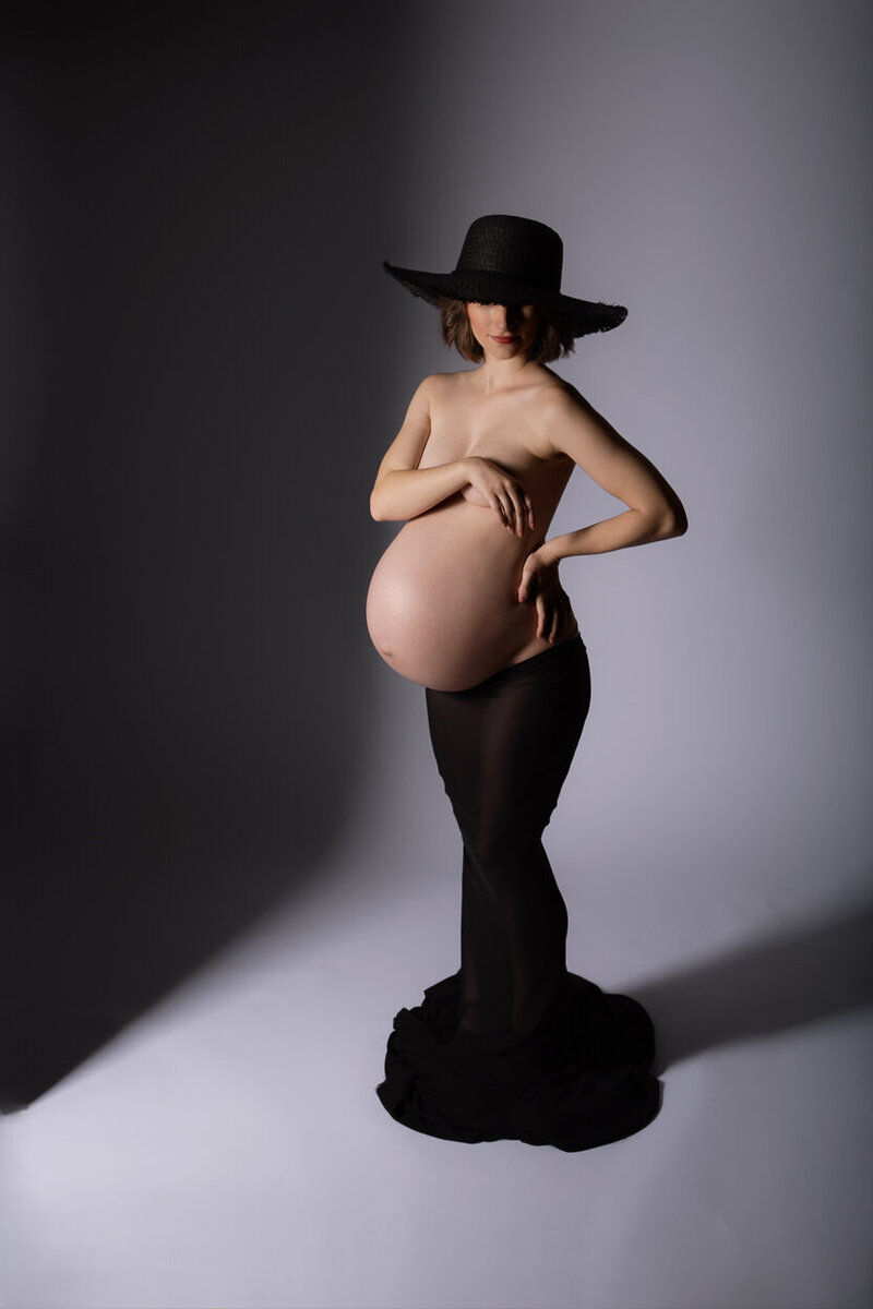 Charlotte mom to be posing with modern lighting showing off her pregnant belly wearing a black hat and skirt. Created in studio by Insley photography