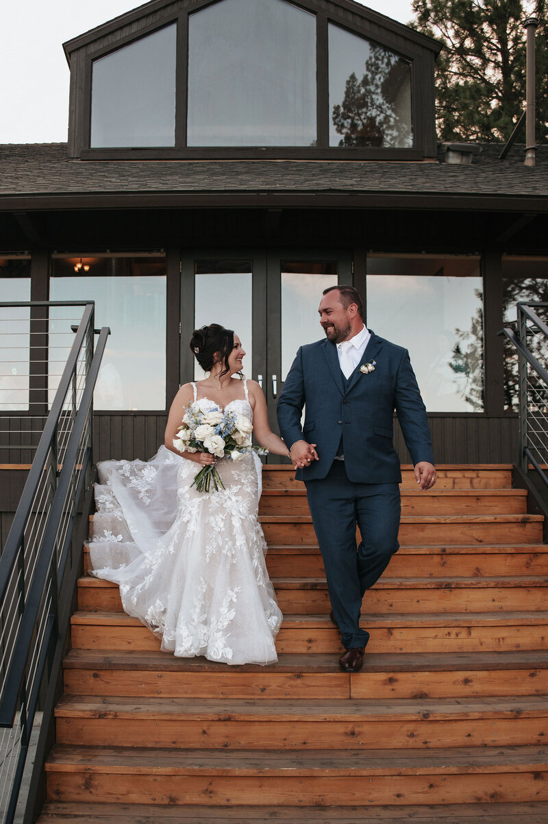 Bridal couple walking together down rustic wooden steps with a modern building in the background, smiling in formal wedding attire at River Run Lodge near Redmond, Oregon.