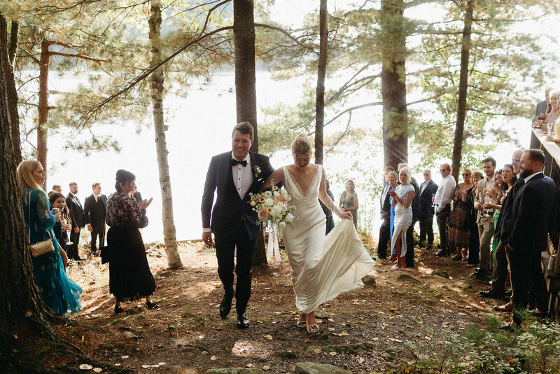 Bride and groom walk hand in hand through a wooded area, surrounded by guests at their Muskoka cottage wedding, as Ontario destination wedding photographer, Jennifer van Son captures every cherished moment.
