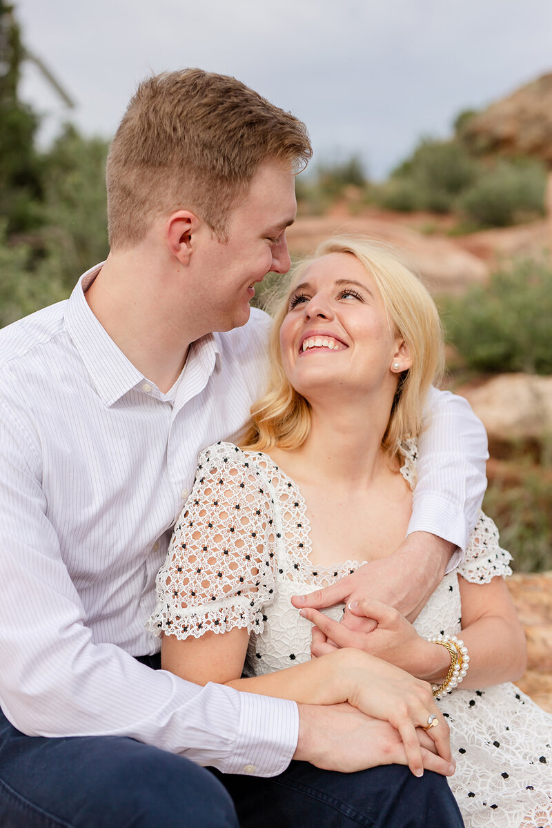 Woman and man hugging and looking at each other , sitting on a red rock at Red Rock Park in Morrison CO. They are posing for an engagement photo session. 