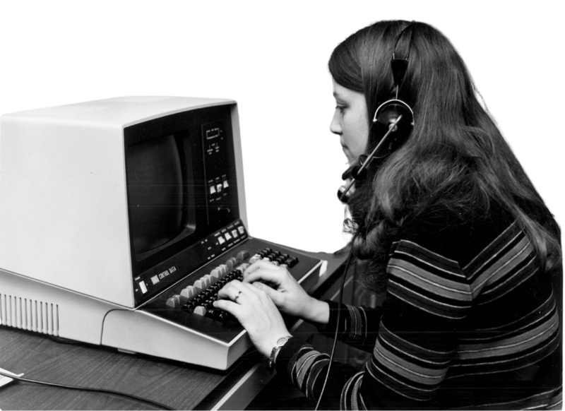 A woman wearing headphones types on a vintage computer terminal with a small screen and keyboard. She appears focused and engaged in her work.