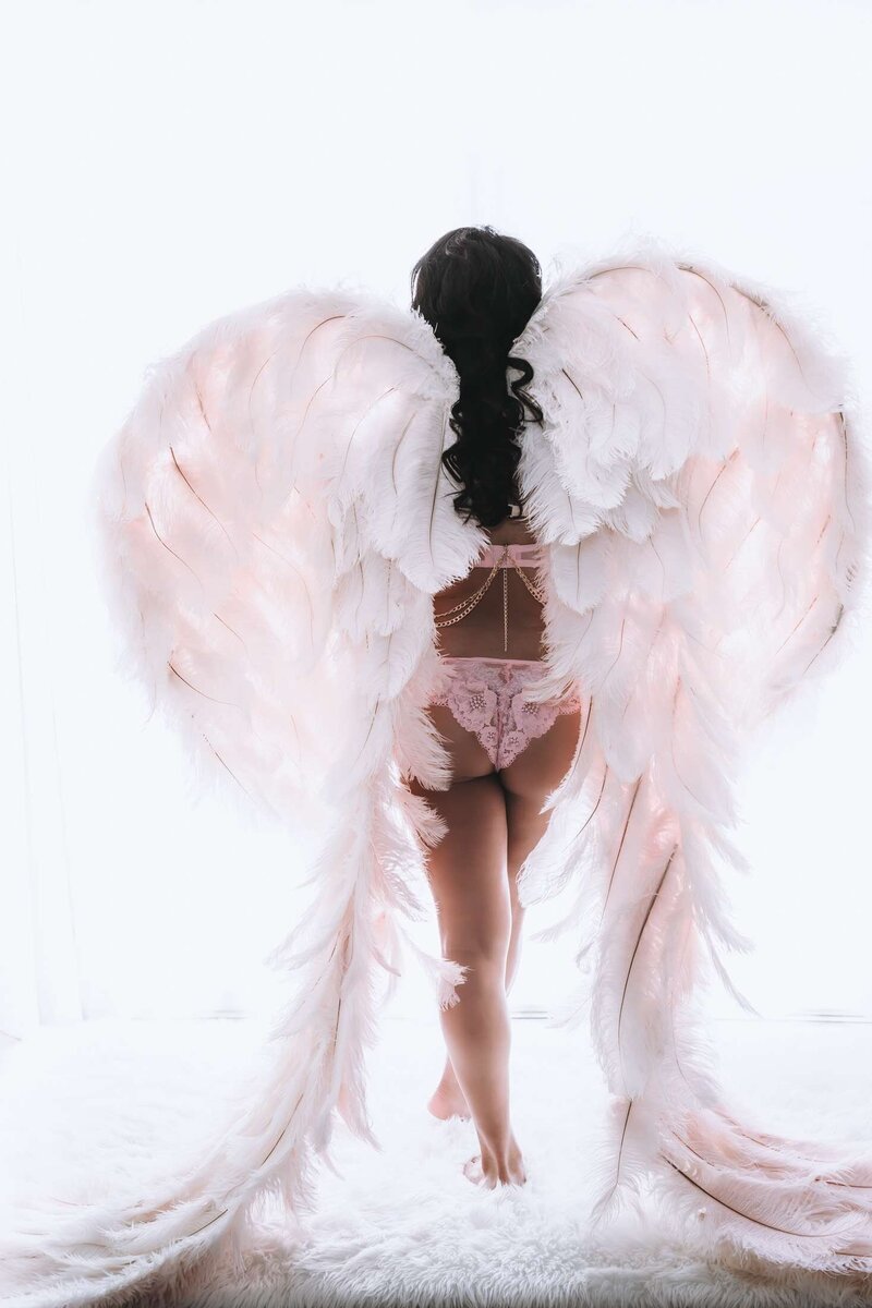 Wearing pink angel wings a woman looks at a bright light during a boudoir photoshoot in Vancouver