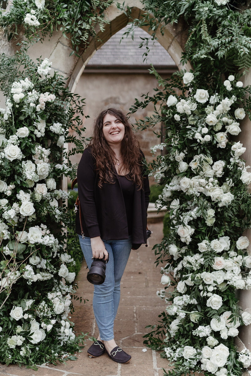 Portland Wedding Photographer Lindsey poses in front of a wedding floral archway in a stone courtyard