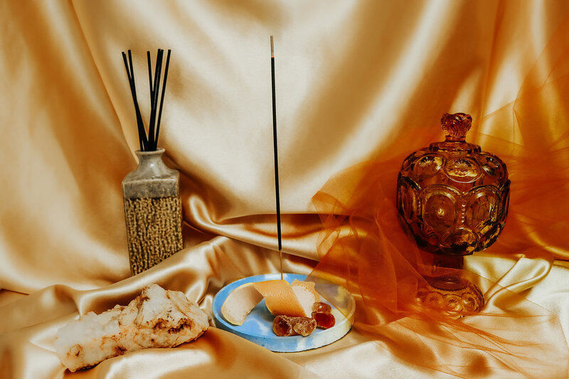 A still life centering a single, lit stick of charcoal incense. The background is a shiny and satiny golden silk. Around the incense in its burner is a clay jar full of unlit charcoal incense, a fresh grapefruit peel, a cool looking rock, and an amber-colored antique glass jar with a lid.