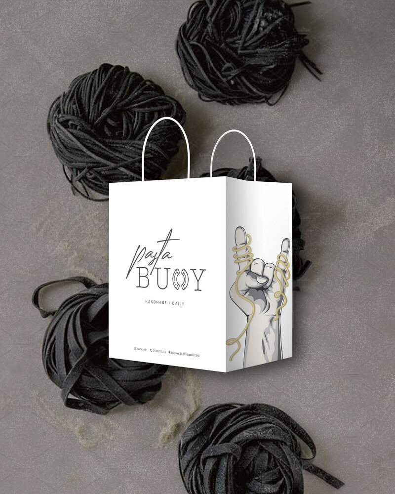 A Pasta Buoy branded shopping bag, featuring the brand’s logo with a hand gesturing with intertwined pasta. The bag is placed beside black coiled pasta, emphasizing the artisanal and unique qualities of the brand.