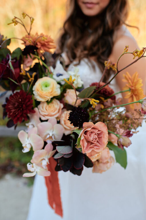 A bride holds a large floral bouquet filled with peach roses, blush orchids, yellow kangaroos tail, burgundy dahlias and other autumn-inspired flowers at her wedding at Le Belvedere venue in Wakefield Quebec.