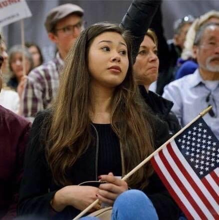 Doris sits among a diverse crowd of individuals behind and beside her, holding an American flag.