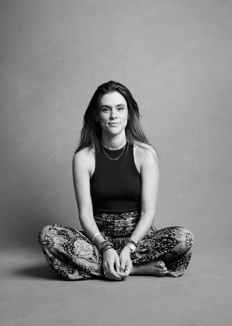Black and white portrait of a woman sitting cross-legged on the floor, looking confidently at the camera. She wears a black sleeveless top and patterned pants, accessorized with layered necklaces and bracelets. A natural, unretouched moment from 'THIS IS ME – A Photography Experience by Katie Campbell' in Atlanta, GA, celebrating authenticity and self-empowerment."