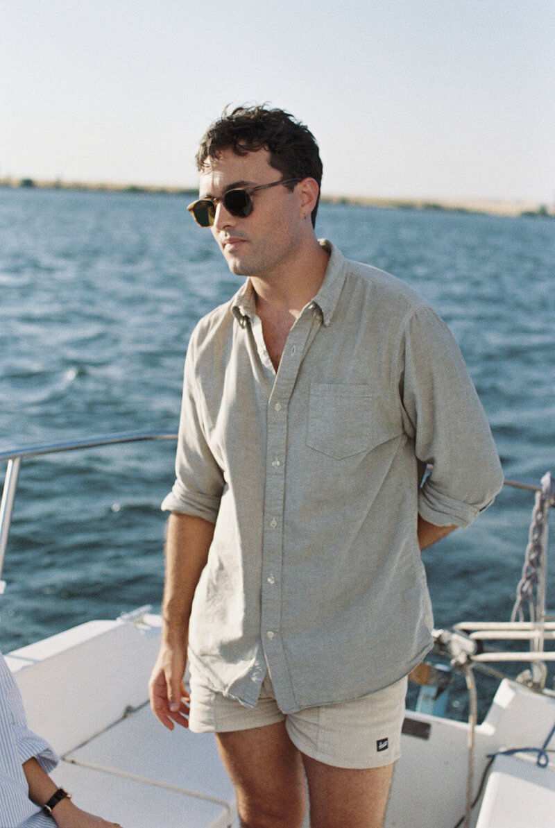 A person in sunglasses and a light shirt stands on a boat with their hands behind their back.
