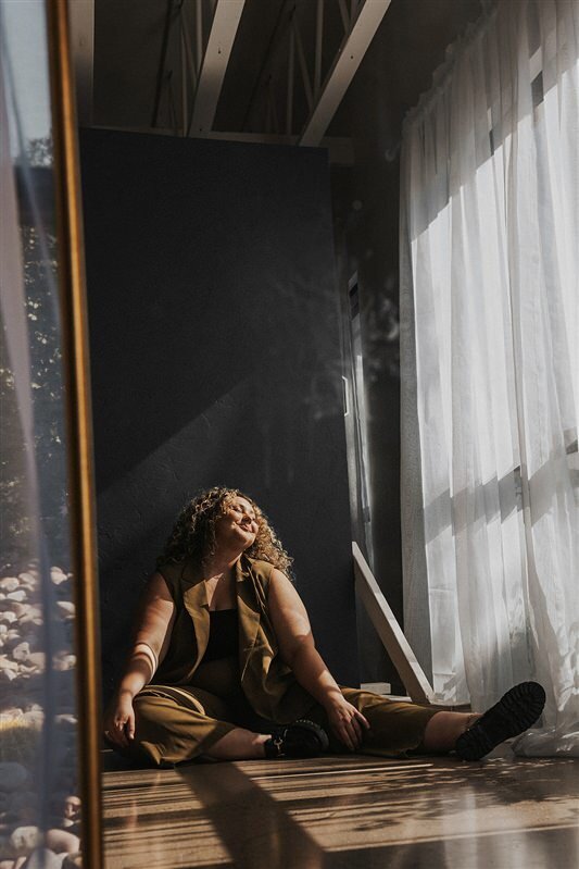 Andi sits on the floor by a large window with sheer white curtains, basking in the sunlight streaming in.