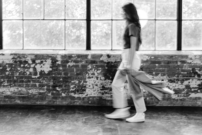 Black-and-white photo of a woman with long hair walking indoors next to a large window. The wall has a weathered brick texture and the floor is plain. The person is in motion and slightly blurred, evoking the dynamic nature of brand identity services they offer.