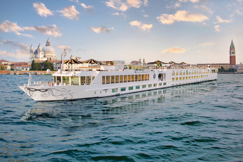 A white river cruise ship sails on calm water near a historic European cityscape with domed buildings and warm, late-day sunlight reflecting on the surface.