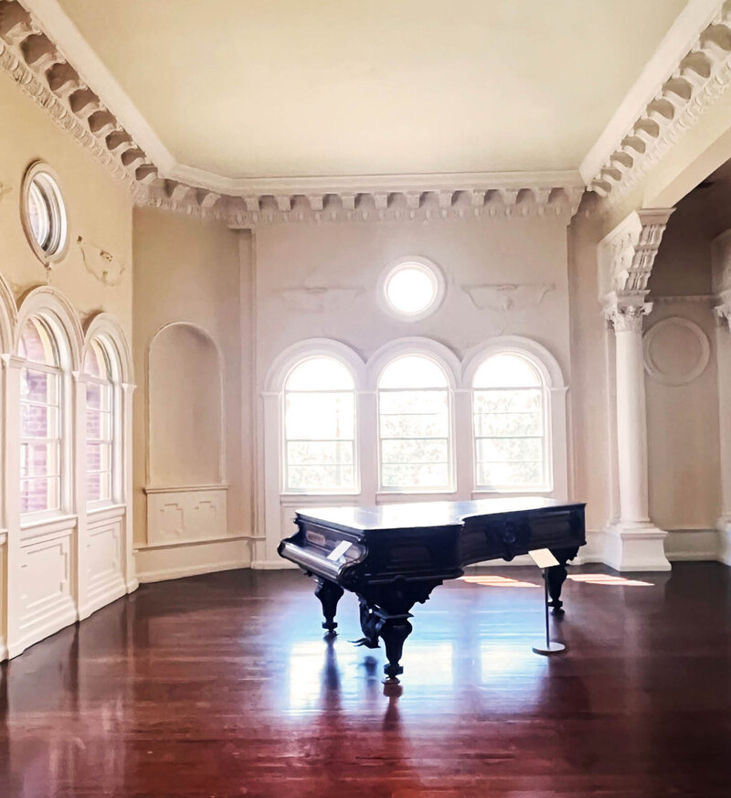 Inside the Lightner Museum in St. Augustine: plaster ceiling moldings, arched and round windows, Greek columns, and a grand piano in a historic architectural setting.