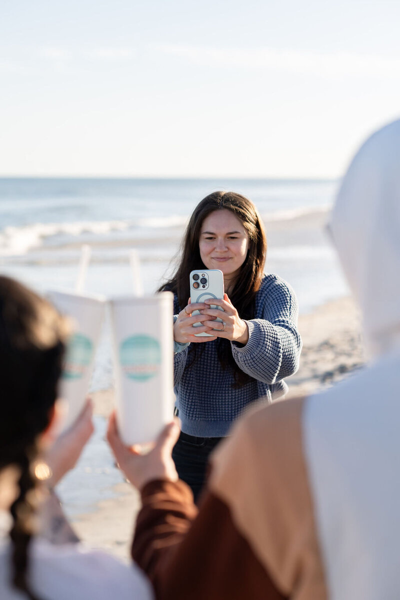 Annie takes a photo of two people holding up their white tumblers using her white phone