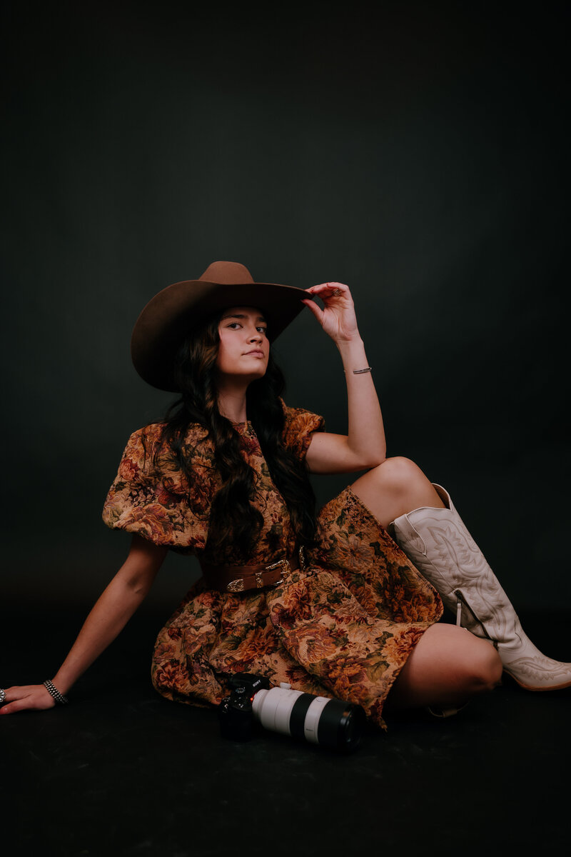 woman sitting on the ground wearing a cowgirl hat and boots with her camera next to her