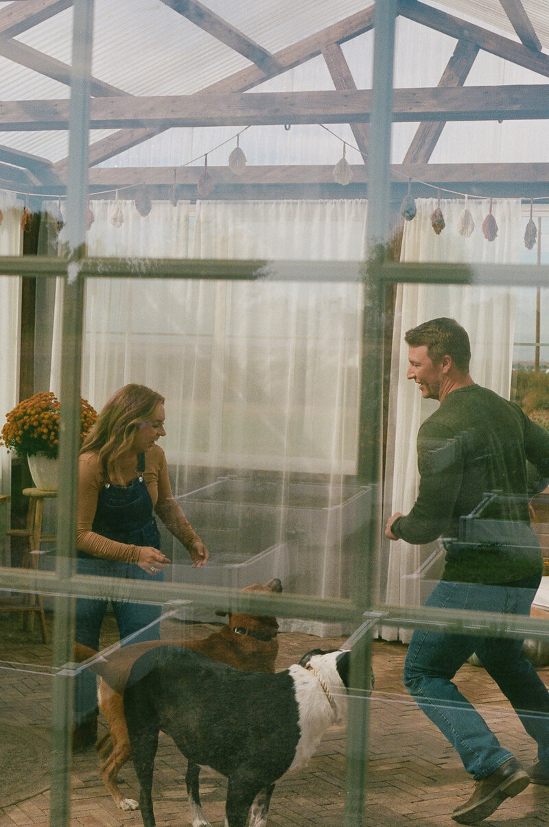 Logan Ferree, a Colorado and destination wedding photography, candidly playing with her dogs and husband in a greenhouse