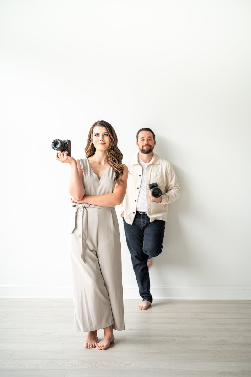 Kala, who is holding her camera in one hand, is standing a few feet in front of Reg. Reg is also holding his camera in one hand with his foot against the wall.