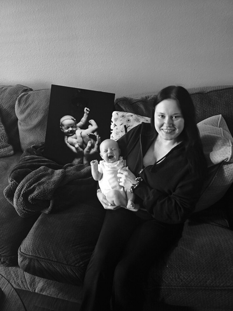 A wall art mockup created by Mobile, AL family photographer. The mock up shows a modern, grey living room with a couch and a coffee table. The wall art showcases photos from a fine art newborn session. One is of a new mother, posing with her newborn on her chest. The baby is sleeping peacefully, skin to skin with his mom. The other photo is a detailed family photo of the baby being held against his dad's chest, while wrapped in a white swaddle with his feet showing. His mom leans in close to the baby and dad, as they all close their eyes to soak in the smallness of their child for an intimate newborn photo.
