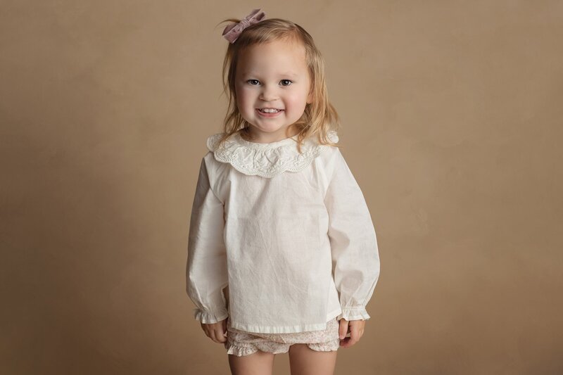 A young toddler girl in a white shirt stands smiling in a studio