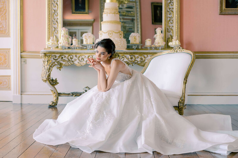 a bride sits on a white chaise longue with her dress billowing around her and an elaborate white and gold wedding cake on display behind her in avington park’s ballroom