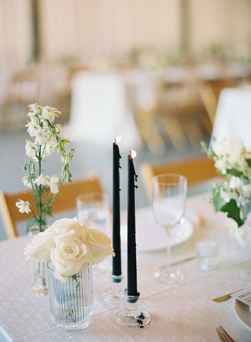 black candles lit next to white roses on a white wedding table in a barn