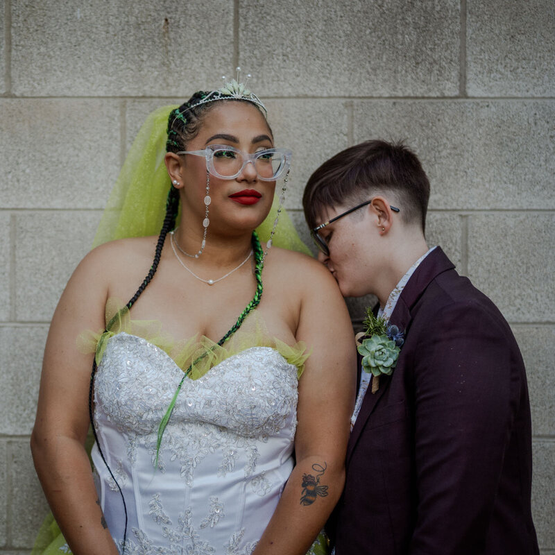 A distinctive and colorful wedding portrait of two brides against a gray brick wall. One bride wears glasses, a white gown with green accents, and a tiara, while the other sports a short haircut and a dark suit. Their tender pose and unique styles make a bold statement.