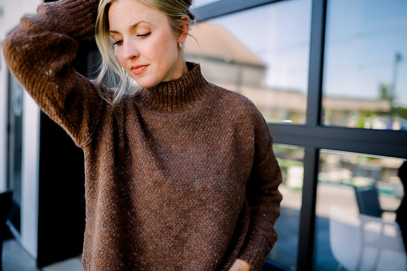 Young female in brown turtleneck sweater outside holding hair up looking down