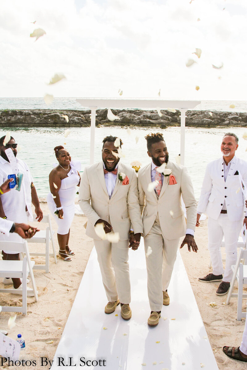 Two men in white suits stroll down a beach aisle, celebrating a destination wedding in a picturesque setting.
