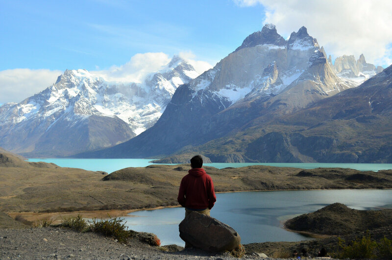 A man in a red shirt sits on a rock looking out at the mountains of Patagonia