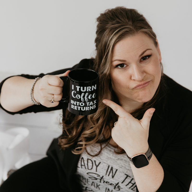 A woman with long, wavy hair holds a black mug that reads "I turn coffee into tax returns" and points at it with a playful expression. She is wearing a casual outfit with a wristwatch, and the background is plain white.