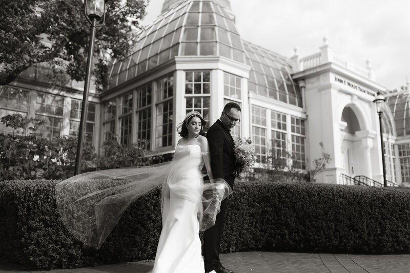 A black and white photo of a bride and groom walking outside a grand glass conservatory, the bride’s veil flowing in the wind.