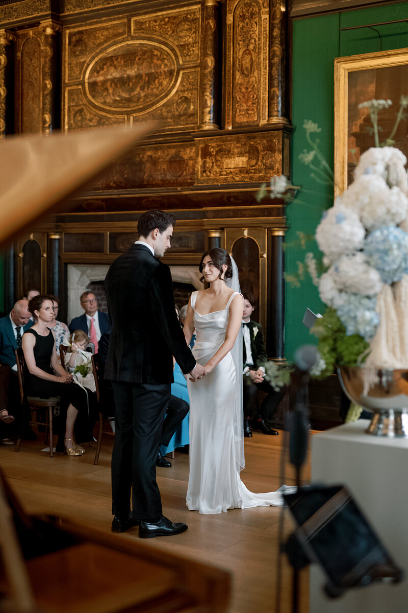 Bride and groom holding hands during emotional wedding ceremony at The Charterhouse, surrounded by floral installations and historic interiors.